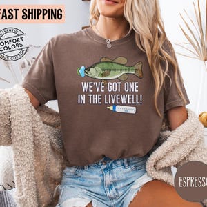 May include: Espresso-colored t-shirt with a graphic of a fish wearing a pacifier and the text "WE'VE GOT ONE IN THE LIVEWELL!". The shirt is made of 100% cotton and features the words "FAST SHIPPING" and "AUTHENTIC COMFORT COLORS".