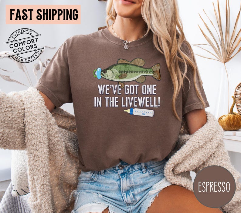 May include: Espresso-colored t-shirt with a graphic of a fish wearing a pacifier and the text "WE'VE GOT ONE IN THE LIVEWELL!". The shirt is made of 100% cotton and features the words "FAST SHIPPING" and "AUTHENTIC COMFORT COLORS".