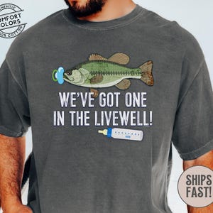 May include: Dark gray t-shirt with a graphic of a fish wearing a pacifier and a baby bottle. Text reads "WE'VE GOT ONE IN THE LIVEWELL!". The shirt is made of 100% cotton.