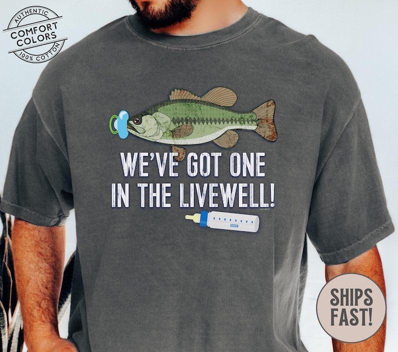 May include: Dark gray t-shirt with a graphic of a fish wearing a pacifier and a baby bottle. Text reads "WE'VE GOT ONE IN THE LIVEWELL!". The shirt is made of 100% cotton.