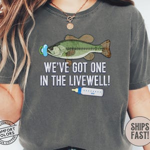 May include: Dark gray t-shirt with a graphic of a fish wearing a pacifier and a baby bottle. The text reads, "WE'VE GOT ONE IN THE LIVEWELL!" The shirt is made of 100% cotton.
