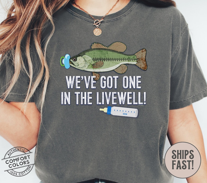 May include: Dark gray t-shirt with a graphic of a fish wearing a pacifier and a baby bottle. The text reads, "WE'VE GOT ONE IN THE LIVEWELL!" The shirt is made of 100% cotton.