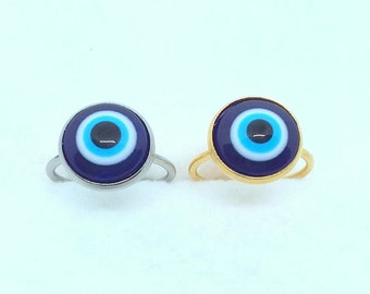 May include: Two rings with an evil eye design. One ring has a silver band, and the other has a gold band. The evil eye design features a dark blue outer circle, a light blue inner circle, and a black center.