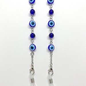 May include: Two eyeglass chains featuring blue and white evil eye beads and dark blue beads. The chains are silver-coloured and have clear rubber loops for attaching to glasses. The chains are displayed against a white background.
