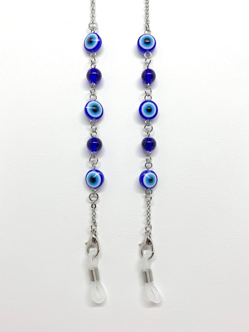 May include: Two eyeglass chains featuring blue and white evil eye beads and dark blue beads. The chains are silver-coloured and have clear rubber loops for attaching to glasses. The chains are displayed against a white background.