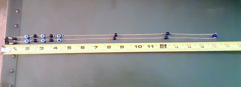 May include: A silver chain necklace featuring blue and white evil eye beads. The necklace is about 43 cm long, measured with a yellow tape measure. The beads are dark and light blue, with an eye design in the center.