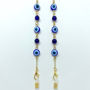 May include: Gold-coloured chain glasses holder featuring blue and white evil eye beads and dark blue beads. The chain has gold-coloured clasps and clear rubber loops for attaching to glasses. The glasses chain is against a white background.