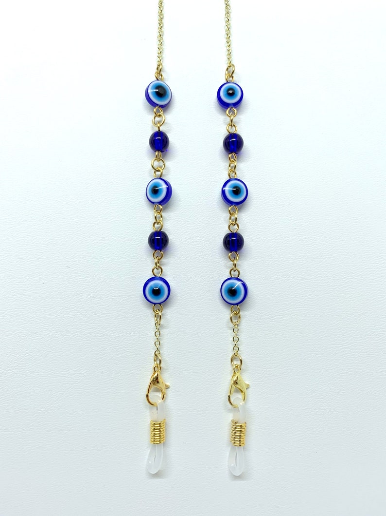 May include: Gold-coloured chain glasses holder featuring blue and white evil eye beads and dark blue beads. The chain has gold-coloured clasps and clear rubber loops for attaching to glasses. The glasses chain is against a white background.