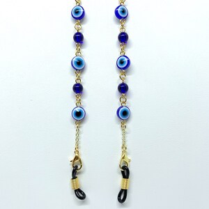 May include: Gold-coloured eyeglass chain featuring blue and white evil eye beads and dark blue beads. The chain has gold-coloured clasps and black rubber loops for attaching to glasses. The chain is displayed against a white background.