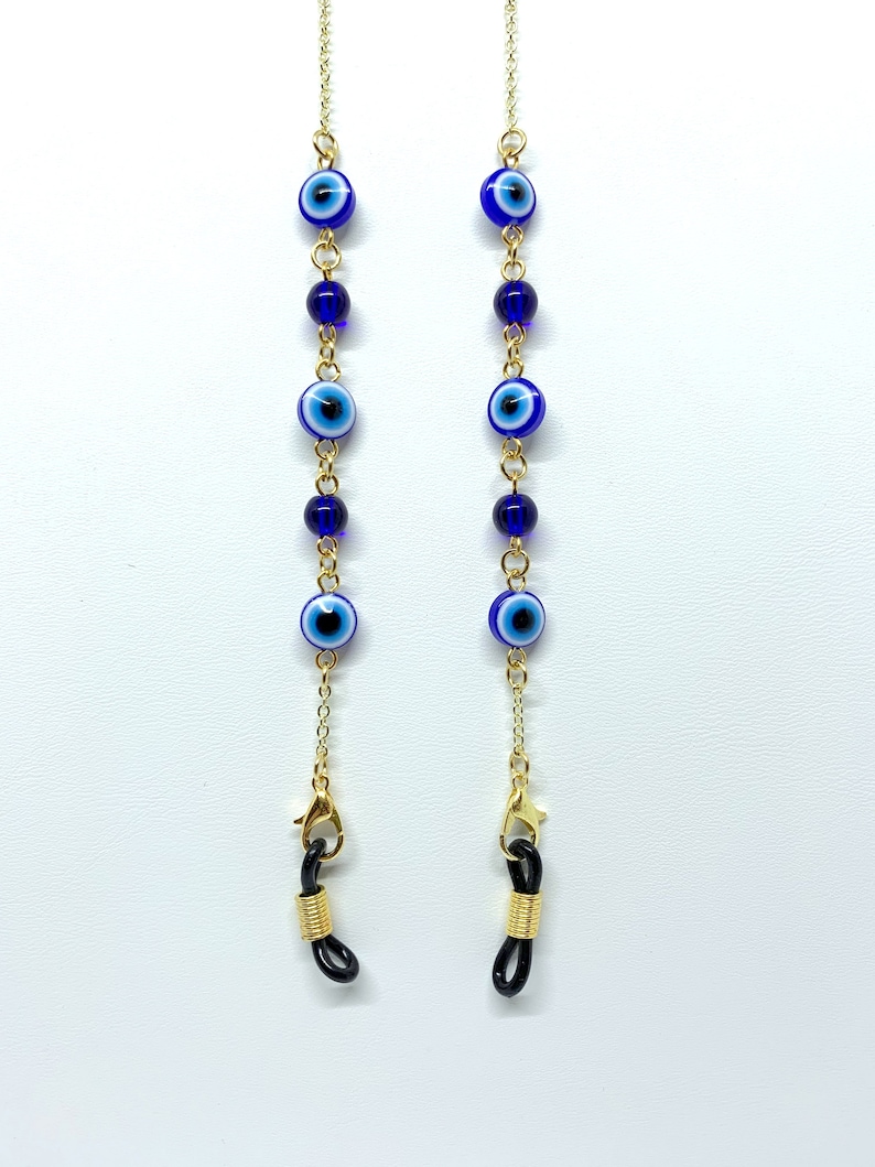 May include: Gold-coloured eyeglass chain featuring blue and white evil eye beads and dark blue beads. The chain has gold-coloured clasps and black rubber loops for attaching to glasses. The chain is displayed against a white background.