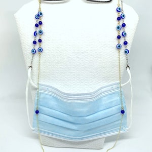 May include: A gold-coloured chain with blue evil eye beads is draped over a light blue face mask and black spectacles. The chain is designed to hold the glasses and mask, with a white background.