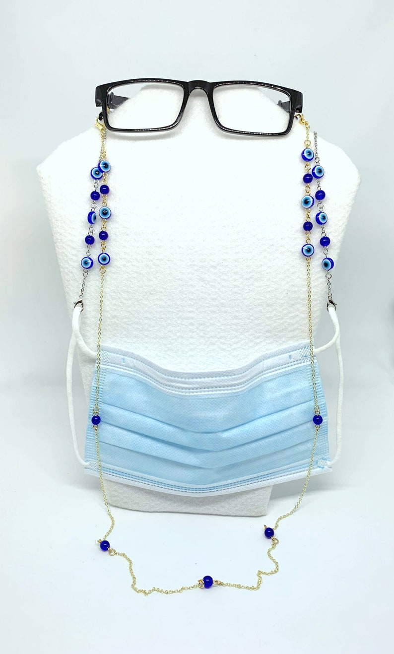 May include: A gold-coloured chain with blue evil eye beads is draped over a light blue face mask and black spectacles. The chain is designed to hold the glasses and mask, with a white background.