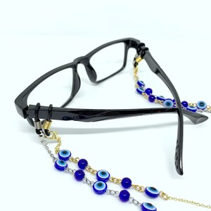 May include: Black-framed spectacles with a decorative chain. The chain features blue and white evil eye beads and is made of gold and silver-coloured metal links. The glasses are on a white background.