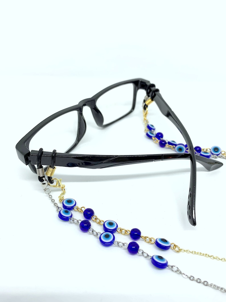 May include: Black-framed spectacles with a decorative chain. The chain features blue and white evil eye beads and is made of gold and silver-coloured metal links. The glasses are on a white background.
