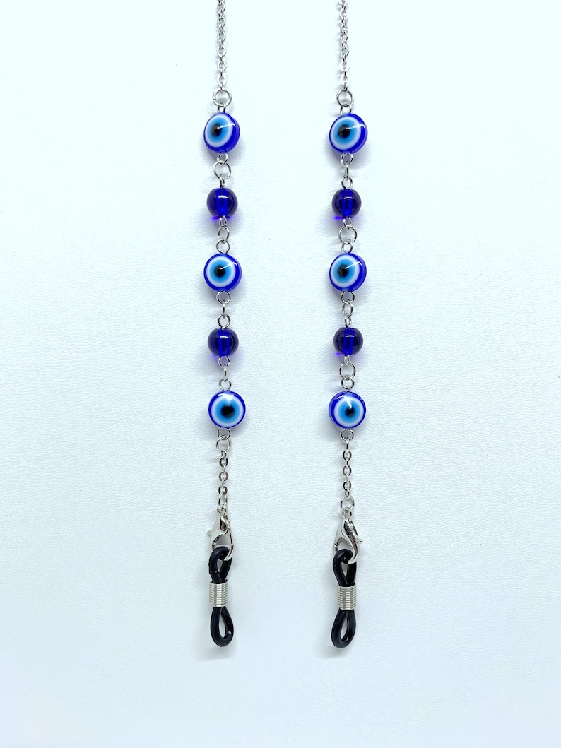 May include: Two eyeglass chains featuring blue and white evil eye beads and dark blue beads. The chains are silver-coloured and have small clasps for attaching to glasses. The background is white.
