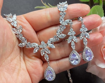 May include: A silver-toned jewelry set featuring a necklace and matching earrings. The necklace and earrings have a floral design with clear, sparkling stones and pear-shaped, light purple stones. The set is displayed against a dark background with a pink rose.