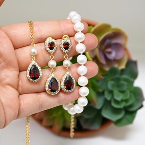 May include: A set of gold earrings with red gemstone teardrop pendants and a matching gold necklace with a single pearl pendant. The necklace is made of white pearls.