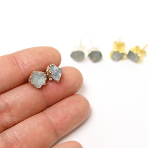 May include: A pair of earrings with a raw, light blue gemstone set in a gold setting. The earrings are shown on a hand.