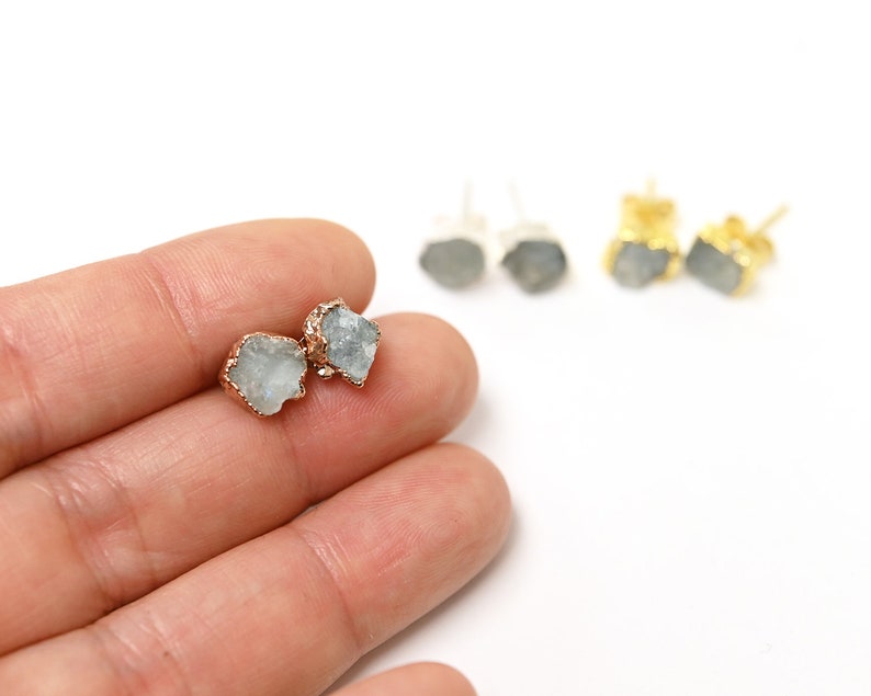 May include: A pair of earrings with a raw, light blue gemstone set in a gold setting. The earrings are shown on a hand.