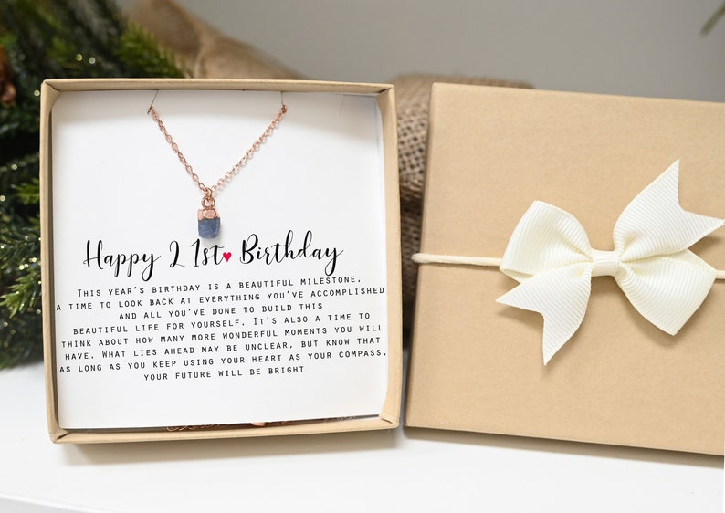 May include: A gift box with a white card that reads "Happy 21st Birthday". The card is inside a brown box with a white bow. The card has a message about looking back at accomplishments and building a beautiful life. The box also contains a necklace with a small blue stone pendant.