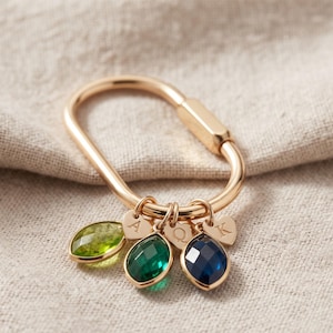 May include: A gold-toned oval carabiner clasp featuring three faceted teardrop-shaped gemstones in light green, dark green, and dark blue, along with three small heart-shaped charms engraved with the letters A, Q, and K respectively.