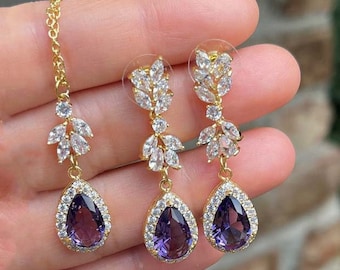 Puede incluir: Un par de pendientes de oro con una piedra púrpura en forma de lágrima y un diseño de hojas. Los pendientes están adornados con piedras de circonita cúbica transparentes.