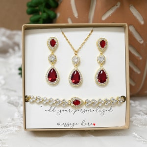 May include: A set of red and gold jewelry in a white box. The set includes a necklace, earrings, and a bracelet. The jewelry features teardrop-shaped red stones surrounded by clear stones. The box has a white card with the text "add your personalized message here".