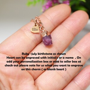 May include: A gold necklace with a small heart charm engraved with the name "Ellie" and a raw ruby pendant. The necklace is shown on a hand.