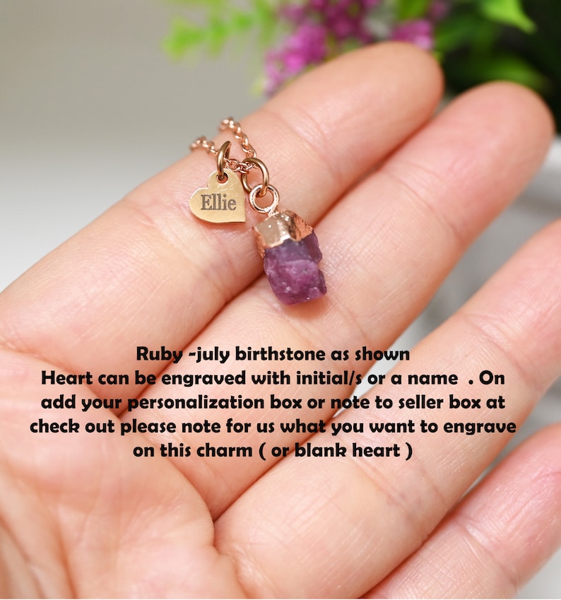 May include: A gold necklace with a small heart charm engraved with the name "Ellie" and a raw ruby pendant. The necklace is shown on a hand.