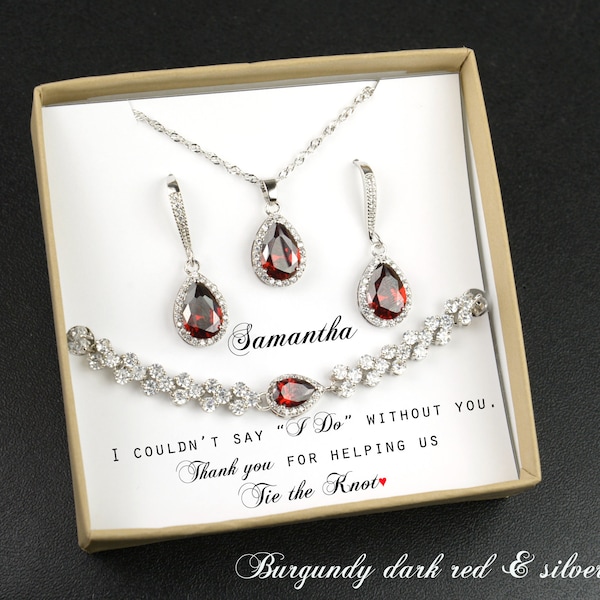 May include: A silver jewellery set with a teardrop pendant necklace, earrings, and bracelet. The teardrop stones are a deep red colour and are surrounded by smaller clear stones. The jewellery is in a box with a message that says "I couldn't say "I Do" without you. Thank you for helping us tie the knot." The box also says "Burgundy dark red & silver".