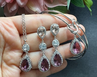 Puede incluir: Un conjunto de joyas que incluye un collar, una pulsera y unos pendientes de plata. El collar y los pendientes tienen un colgante en forma de lágrima con una piedra central transparente y un halo de piedras más pequeñas. La pulsera es una pulsera de plata con un diseño de nudo. Los pendientes tienen una piedra rosa, mientras que el collar y la pulsera tienen una piedra morada.