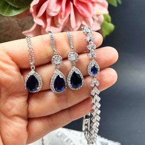 May include: A set of teardrop-shaped earrings, a necklace, and a bracelet. Each piece features a large, dark blue gemstone surrounded by a halo of smaller, clear gemstones. The jewellery is silver-toned and has a delicate, elegant design. navy blue #7