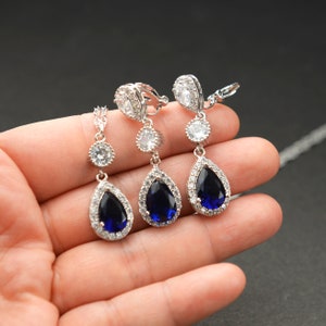 May include: A set of blue sapphire teardrop earrings and a matching necklace. The earrings are clip-on and the necklace has a pendant with a large blue sapphire surrounded by smaller clear stones.