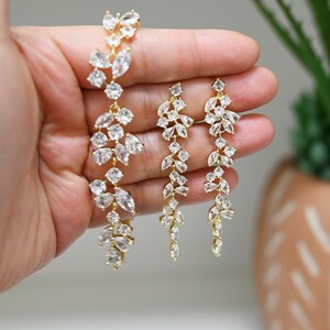 May include: A gold-toned bracelet and matching earrings set. The jewelry features a cascading design of clear, sparkling crystals. The bracelet and earrings are arranged to show the intricate details of the crystal arrangement.