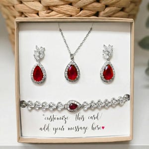 May include: A jewelry set featuring a necklace, earrings, and bracelet with red teardrop-shaped gems. The set includes a silver-toned chain and is presented in a gift box. The text on the card reads "customize this card / add your message here".