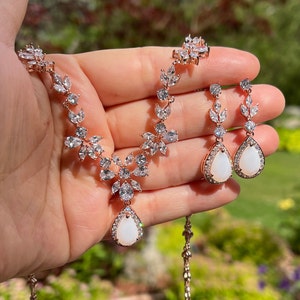 May include: A rose gold bridal necklace and earrings set with a pear-shaped white stone pendant and matching earrings. The necklace features a delicate floral design with clear cubic zirconia stones.