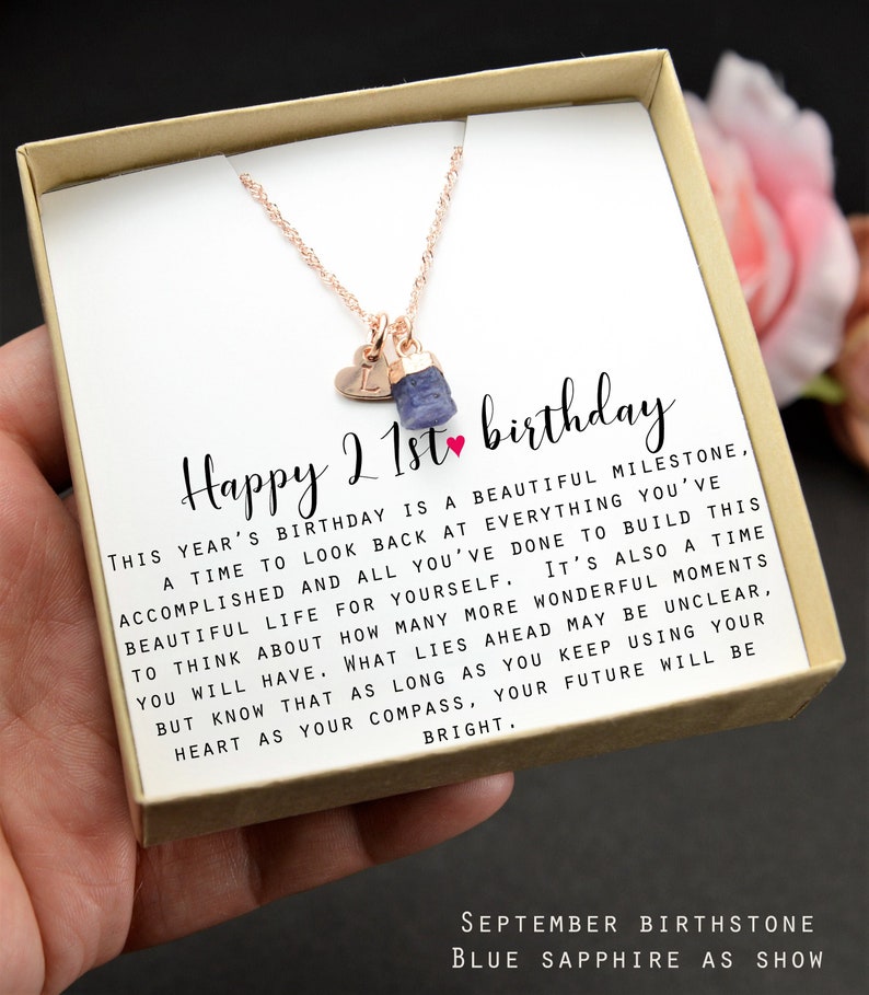 May include: A gift box with a rose gold necklace and a blue sapphire birthstone charm. The box includes a card with the text "Happy 21st birthday" and a message about looking back at accomplishments and building a beautiful life. The card also mentions that the blue sapphire is the September birthstone.