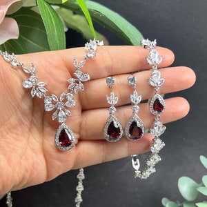 May include: A silver necklace, bracelet, and earrings set with a teardrop-shaped red gemstone surrounded by clear cubic zirconia stones.