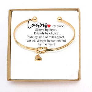May include: Gold bangle bracelet with a knot charm and a heart charm engraved with the name "Lana". The bracelet comes in a box with a message that reads "Cousins by blood. Sisters by heart. Friends by choice. Side by side or miles apart, We will always be connected by the heart."