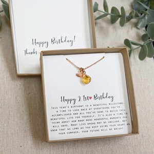 May include: A gift box with a necklace inside. The necklace has a small heart charm and a larger, orange gemstone charm. The box has a card inside that reads "Happy 21st Birthday." This year's birthday is a beautiful milestone. A time to look back at everything you've accomplished and all you've done to build this beautiful life for yourself. It's also a time to think about how many more wonderful moments you will have. What lies ahead may be unclear, but know that as long as you keep using your heart as your compass, your future will be bright.