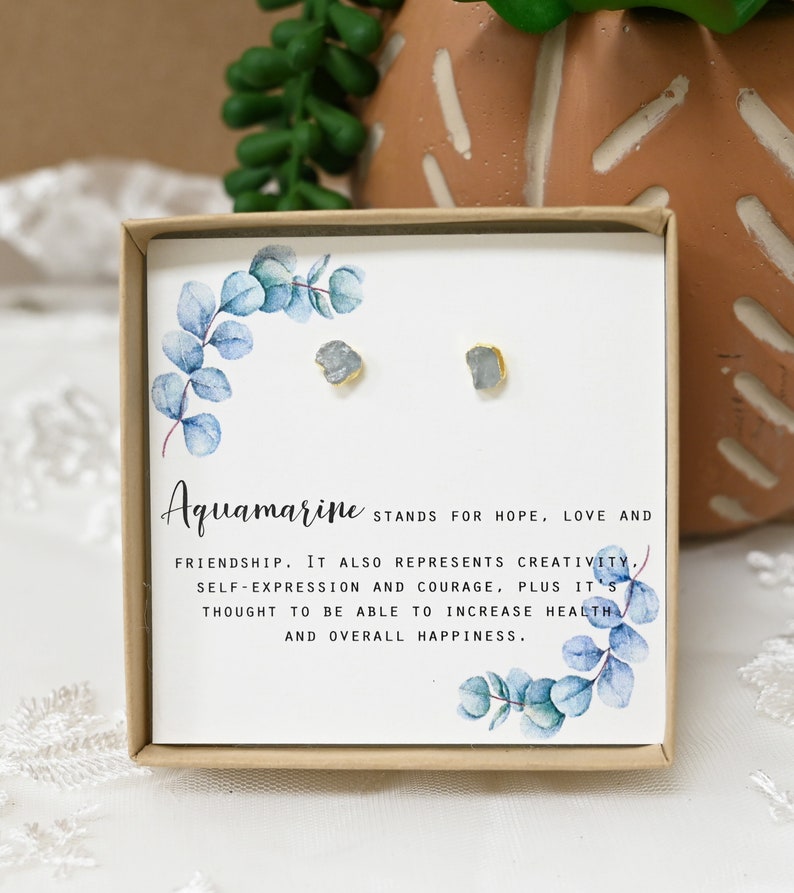 May include: A small box with a pair of aquamarine earrings. The box has a watercolor design of eucalyptus leaves and the text "Aquamarine STANDS FOR HOPE, LOVE AND FRIENDSHIP. IT ALSO REPRESENTS CREATIVITY, SELF-EXPRESSION AND COURAGE, PLUS IT'S THOUGHT TO BE ABLE TO INCREASE HEALTH AND OVERALL HAPPINESS."