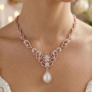 May include: A rose gold necklace with a floral design and a pear-shaped pendant. The necklace features clear crystals and a large, white, pear-shaped faux pearl. The necklace is worn on a person's neck.