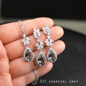 May include: A silver necklace and earrings set with a teardrop-shaped charcoal gray stone surrounded by clear rhinestones. The necklace and earrings feature a delicate leaf design with clear rhinestones.