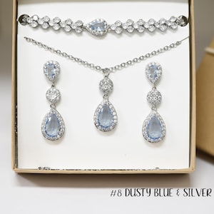 May include: A silver jewelry set with a teardrop-shaped pendant and earrings featuring light blue stones. The set also includes a silver bracelet with clear stones. The jewelry set is in a box with the text "#8 DUSTY BLUE & SILVER".