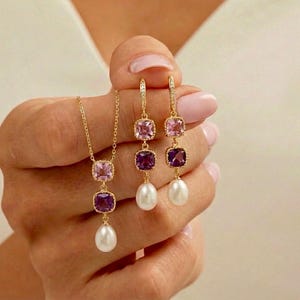 May include: A gold necklace and earrings set featuring square-cut pink and purple gemstones, and white teardrop pearls. The jewelry is held up against a white background.