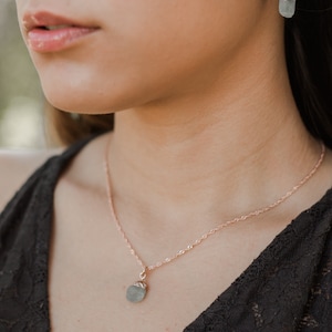 May include: A delicate rose gold necklace with a small, light blue gemstone pendant. The necklace is worn by a person with dark hair.