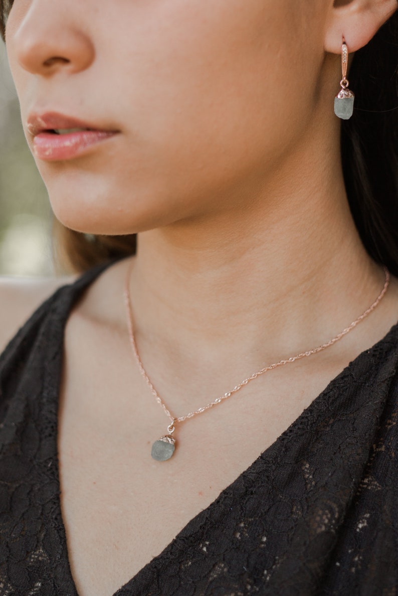 May include: A delicate rose gold necklace with a small, light blue gemstone pendant. The necklace is worn by a person with dark hair.