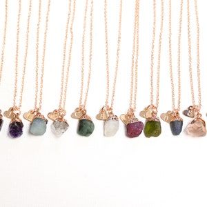 May include: A collection of 12 necklaces with raw gemstone pendants and a small heart charm with an initial. The gemstones are various colors, including red, purple, green, blue, pink, yellow, and brown.