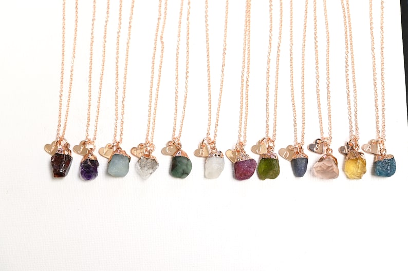 May include: A collection of 12 necklaces with raw gemstone pendants and a small heart charm with an initial. The gemstones are various colors, including red, purple, green, blue, pink, yellow, and brown.
