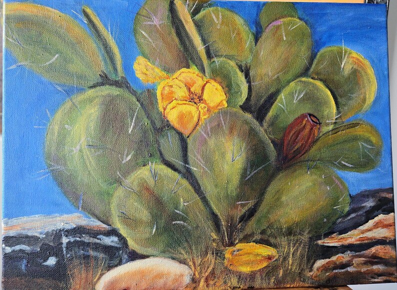 Puede incluir: Una pintura al &oacute;leo sobre lienzo que representa un cactus de nopal con paletas verdes, espinas blancas, una flor amarilla vibrante en plena floraci&oacute;n y un peque&ntilde;o fruto marr&oacute;n rojizo, sobre un cielo azul y terreno rocoso.